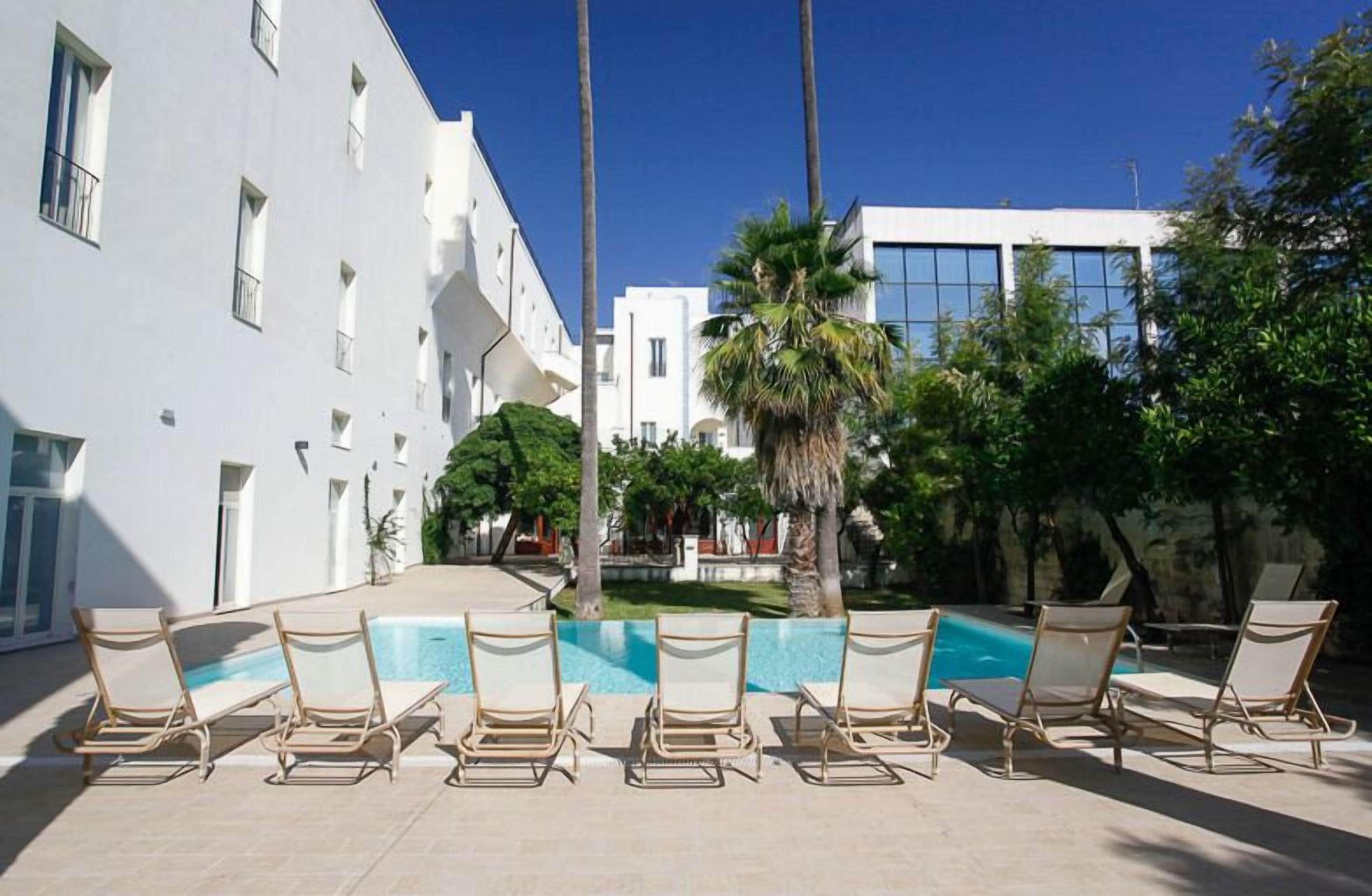 A pool area with lounge chairs and a clear blue pool, surrounded by white buildings and palm trees.