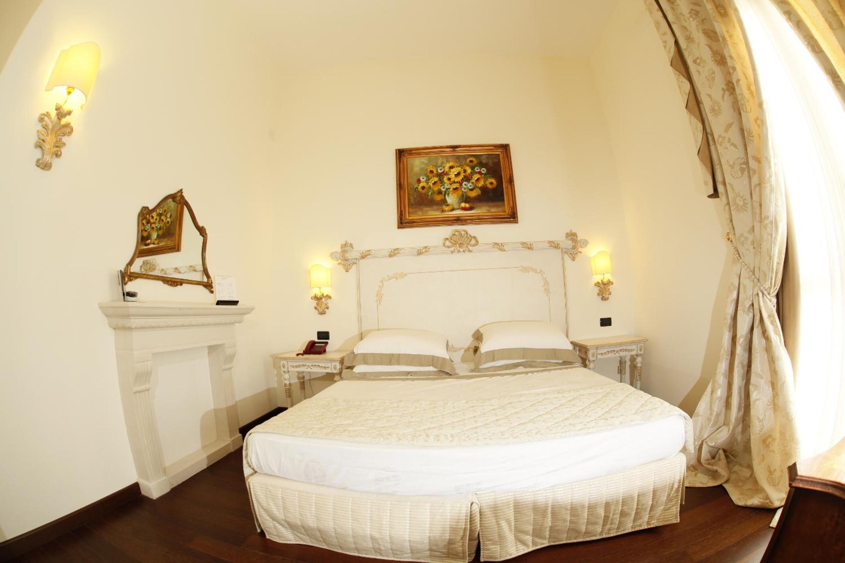 A bedroom with a large bed, a fireplace, a painting of flowers, and a nightstand with a lamp.