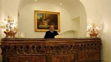A reception desk with a man standing behind it, a framed painting above, and decorative vases on either side.