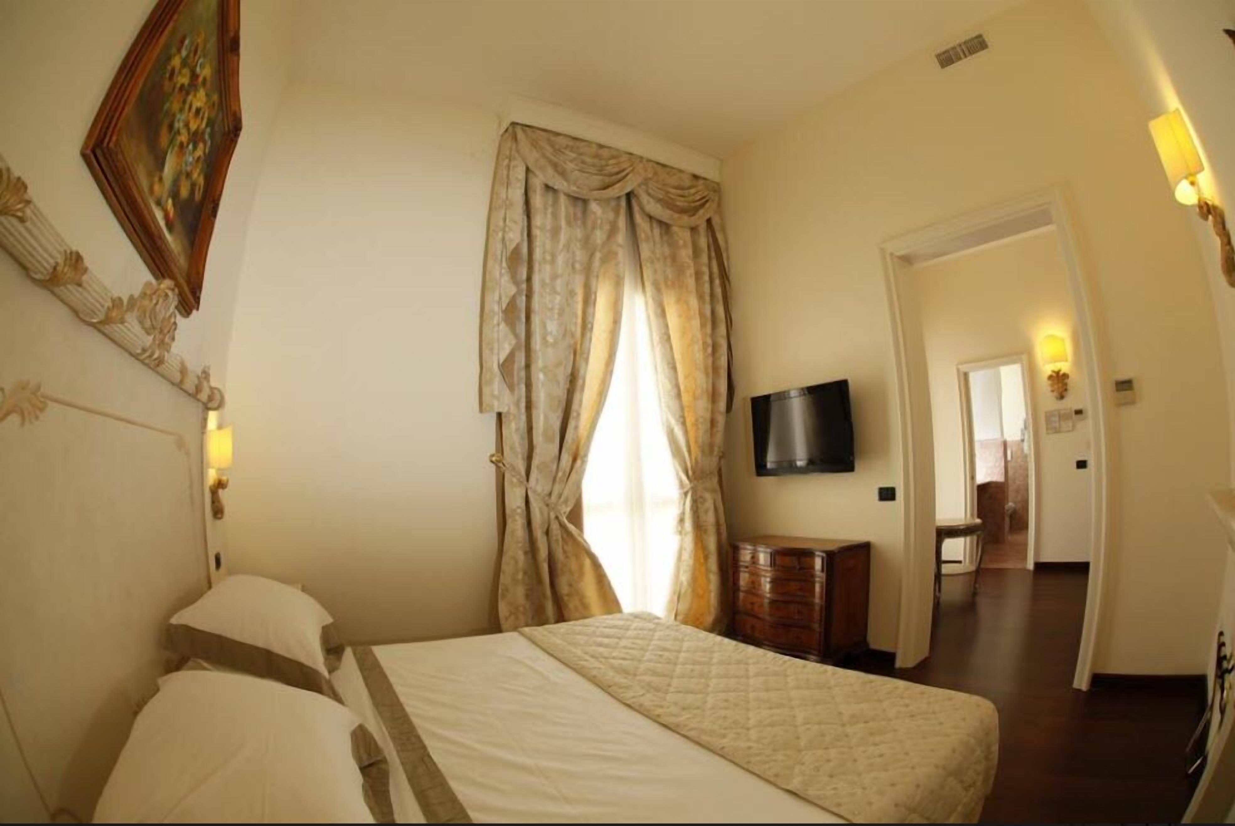 A hotel room with a bed, a TV mounted on the wall, a wooden dresser, and a door leading to another room.