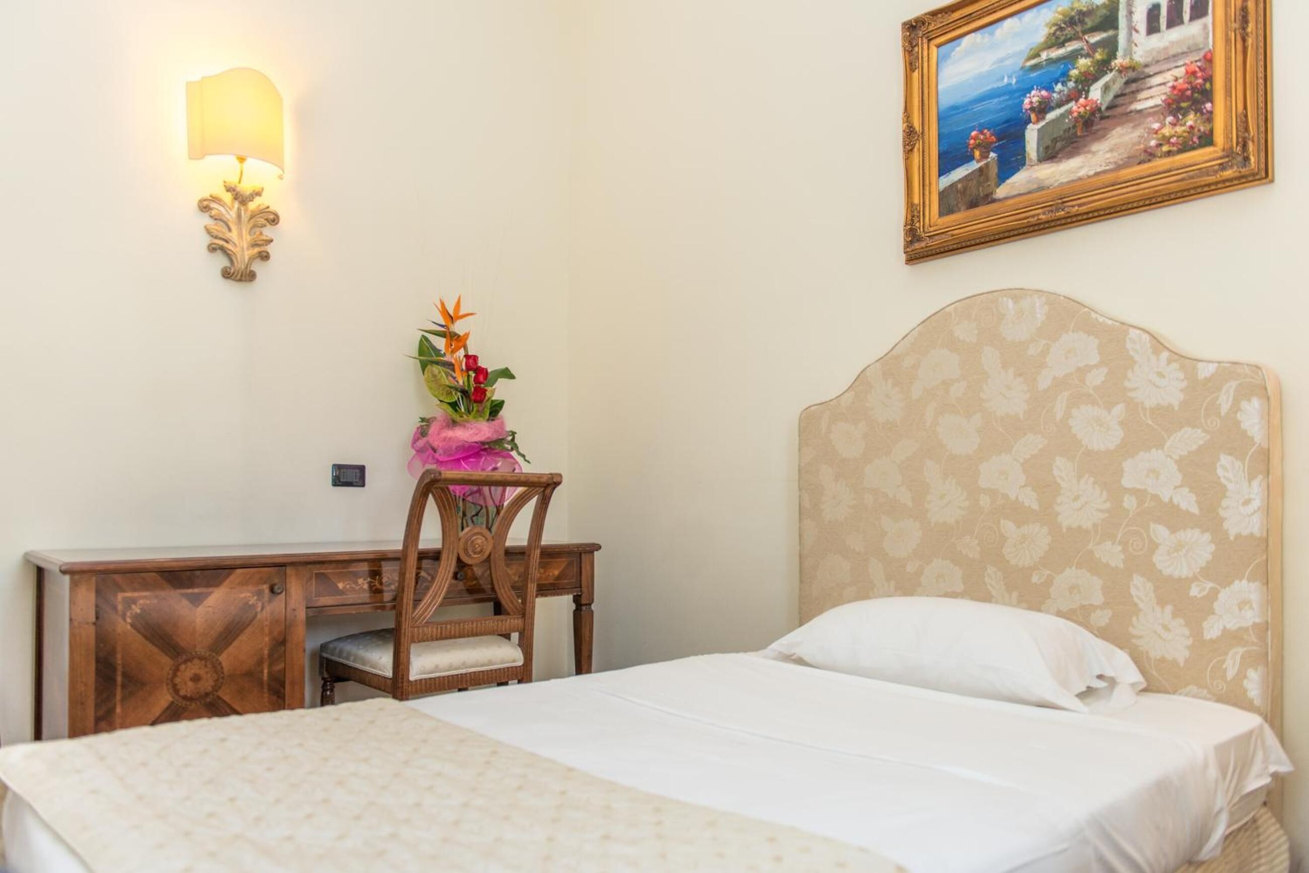 Habitación de hotel con cama, escritorio con silla, lámpara de pared y un cuadro enmarcado.