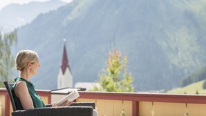 Una persona seduta su una sedia in vimini, che legge un libro, con un paesaggio montano e una chiesa sullo sfondo.