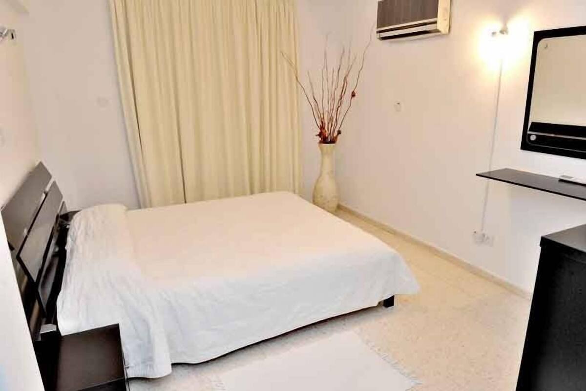 Um quarto com cama, mesa de cabeceira, abajur, vaso com galhos, janela com cortinas e TV na parede.