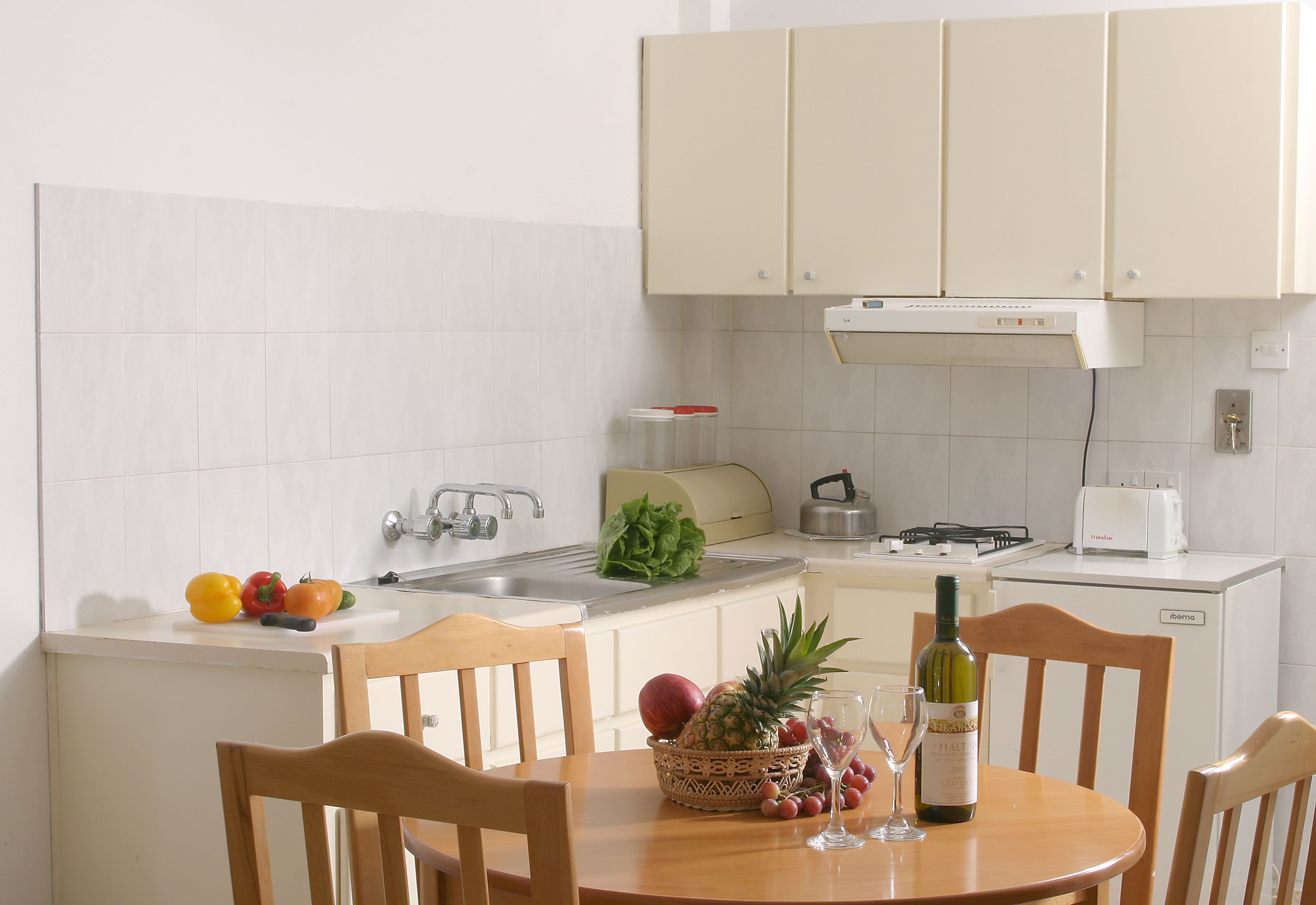 Una cocina con armarios blancos, un lavamanos, una estufa y un tostador. Hay una mesa con sillas, una cesta de fruta y una botella de vino.