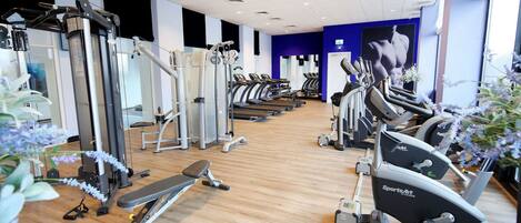 Un gimnasio con diversas máquinas de ejercicio, suelo de madera y pared azul con un espejo grande.