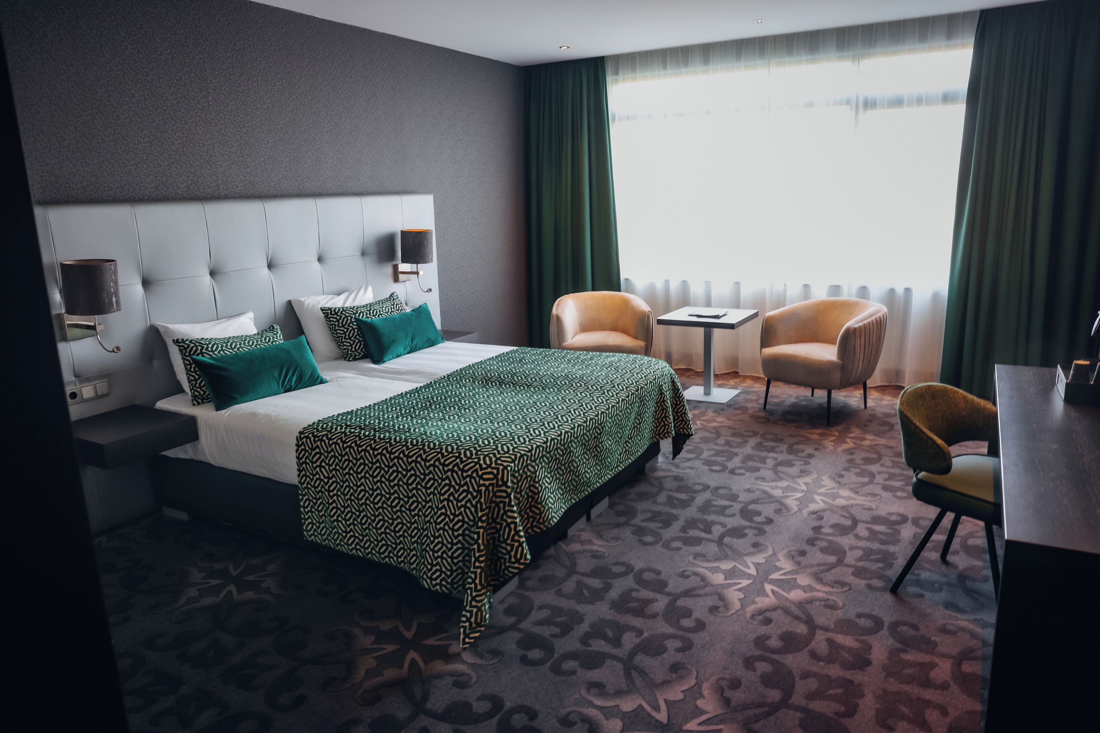 Una habitación de hotel con una cama grande, una mesita de noche, una silla, una pequeña mesa y una ventana con cortinas.