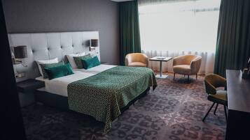 Quarto de hotel com uma cama grande, uma mesa de cabeceira, uma cadeira, uma pequena mesa e uma janela com cortinas.