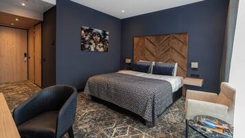 Una habitación de hotel moderna con una cama grande, cabecera con diseño geométrico, zona de estar con una mesa y una obra de arte colgada en la pared.