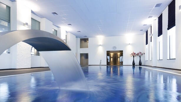 Une vaste piscine intérieure au design moderne, avec une grande structure incurvée en premier plan et une horloge au mur.
