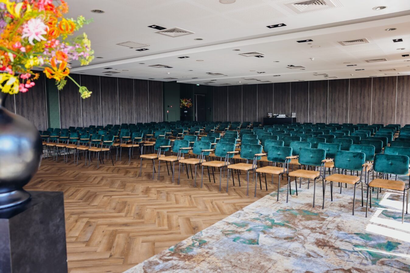A conference room with rows of teal chairs and a wooden floor, a marble table, and a floral arrangement.