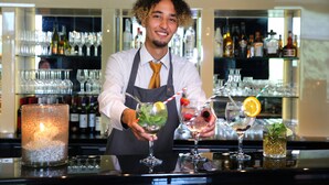 Un barman derrière un comptoir, dans un bar où l’on propose une grande variété de boissons, avec des bouteilles alignées en arrière-plan.