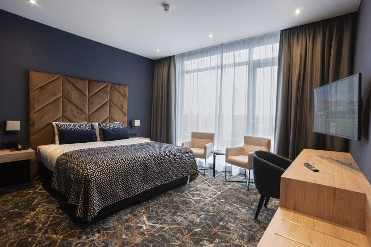 A hotel room with a large bed, a desk, two chairs, a TV, and a view of the city through large windows.