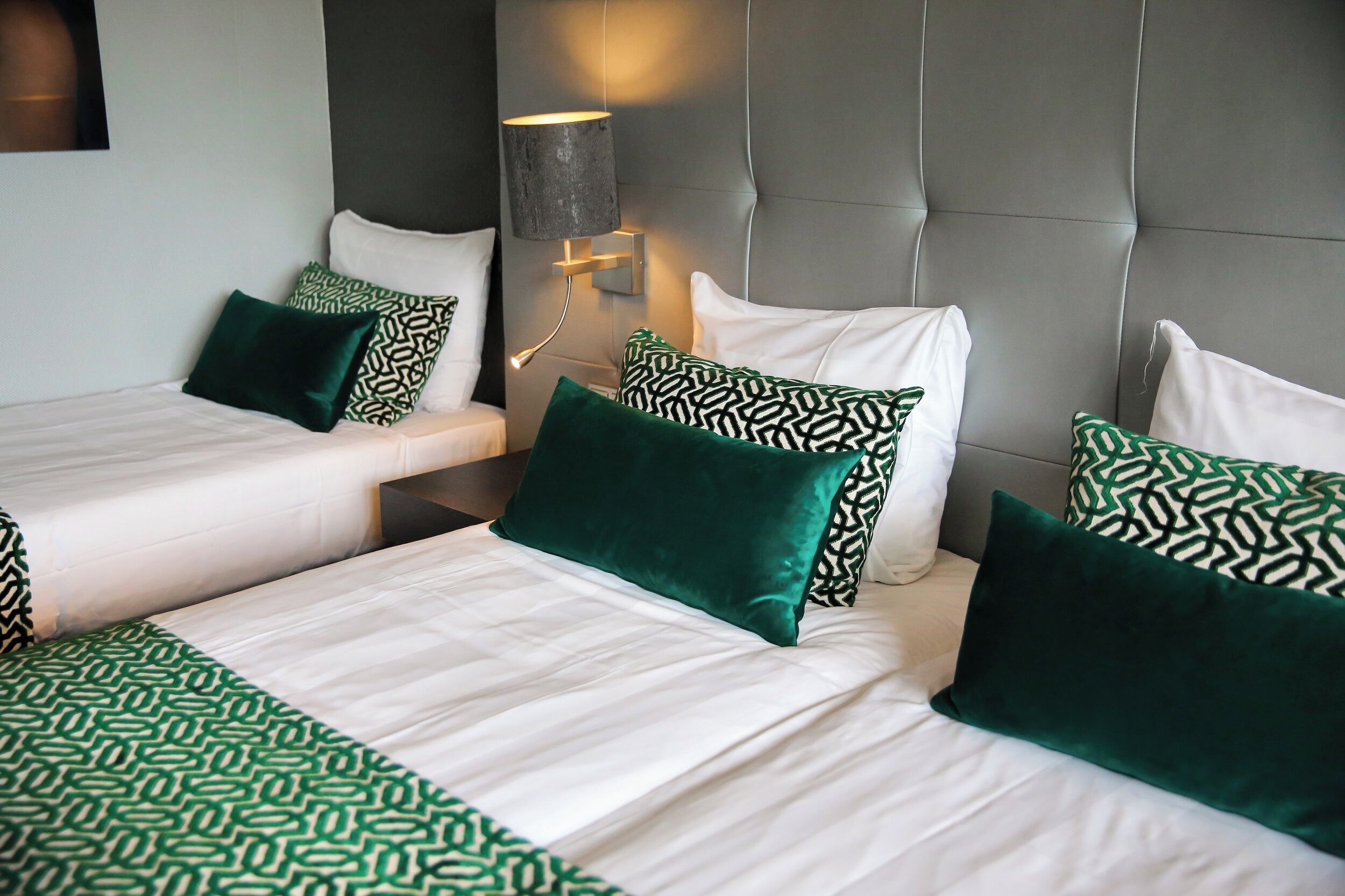 A hotel room with two beds, a headboard, a wall-mounted lamp, and patterned bedding.