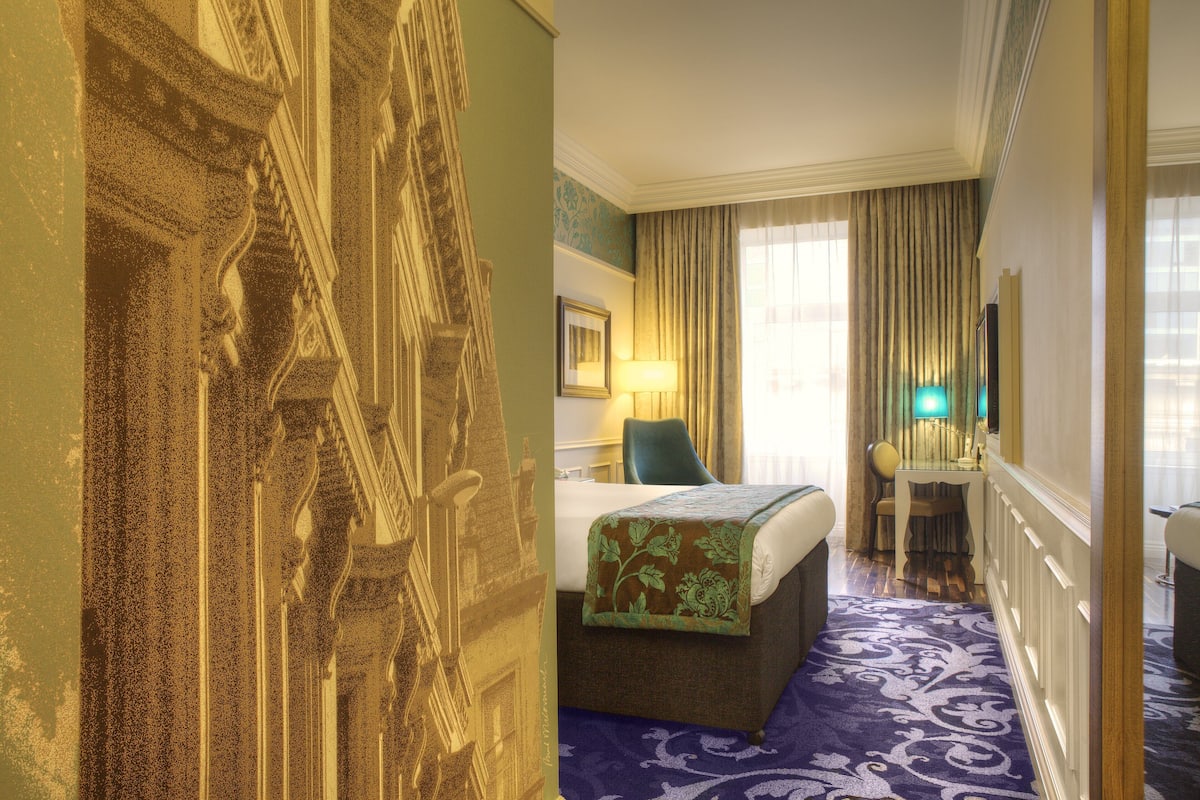 A hotel room with a bed, a desk, a chair, a lamp, and a large window with curtains.