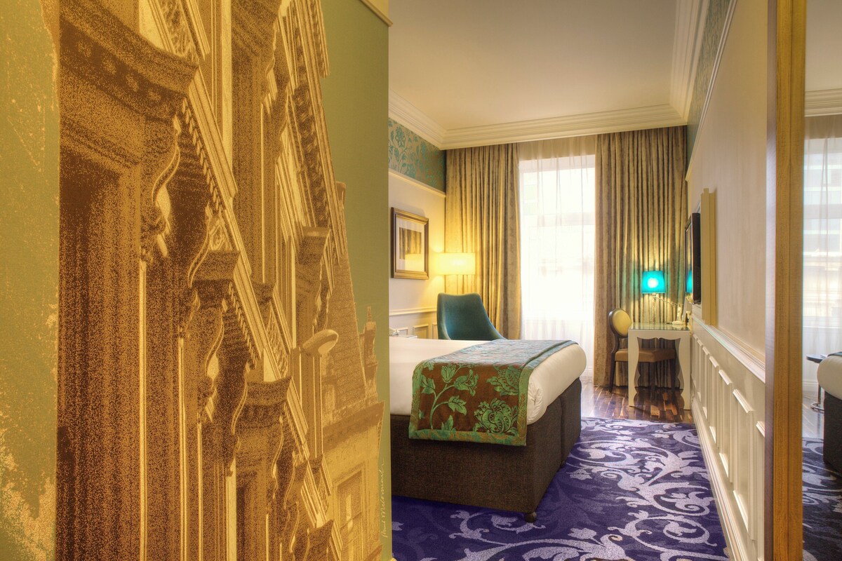 A hotel room with a bed, a desk, a chair, a lamp, and a large window with curtains.