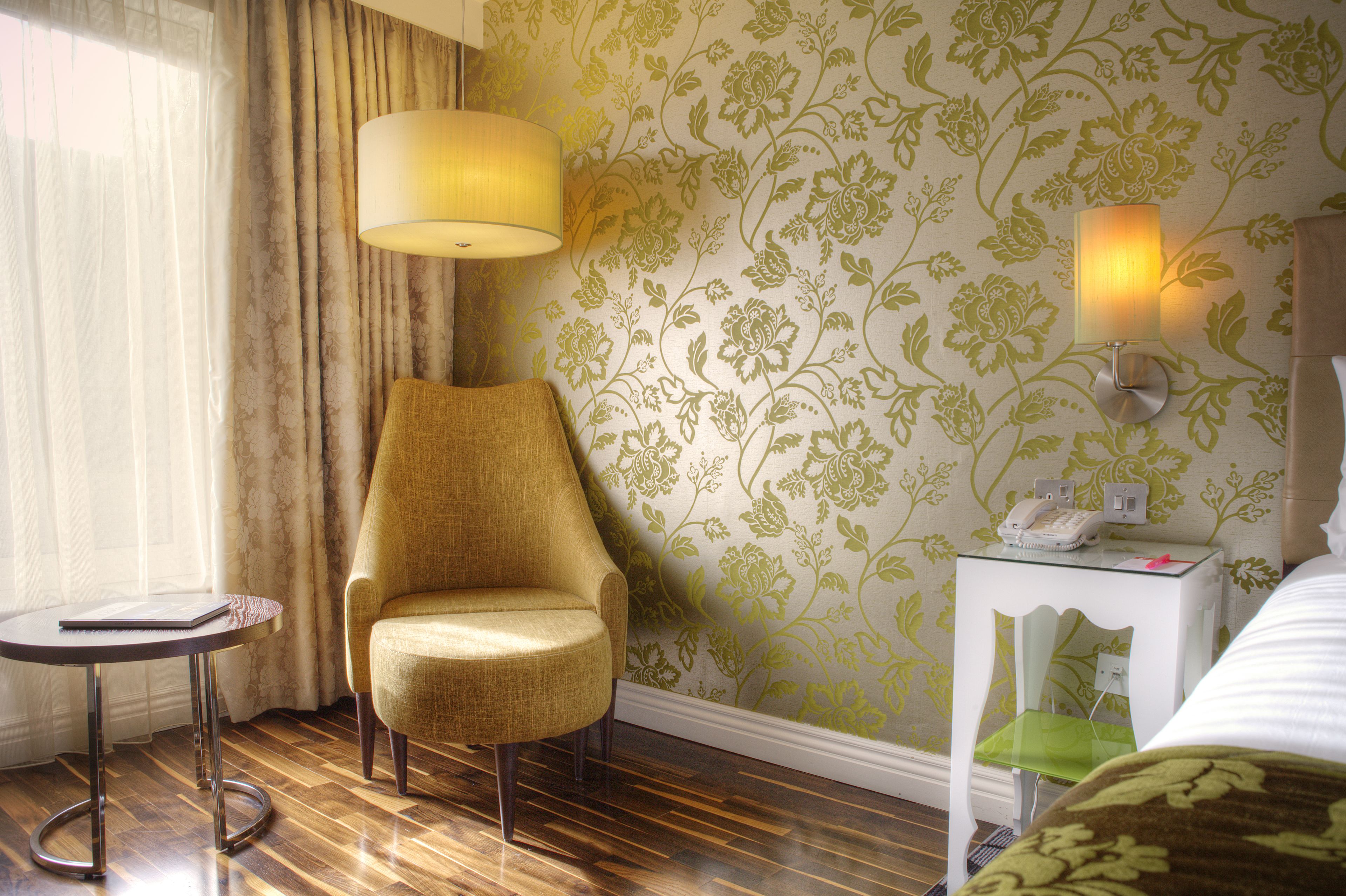 Habitación de hotel con piso de madera, un sillón amarillo, una mesita redonda pequeña, una lámpara, una mesita de noche con un teléfono y papel tapiz floral.