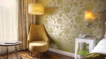 Habitación de hotel con piso de madera, un sillón amarillo, una mesita redonda pequeña, una lámpara, una mesita de noche con un teléfono y papel tapiz floral.