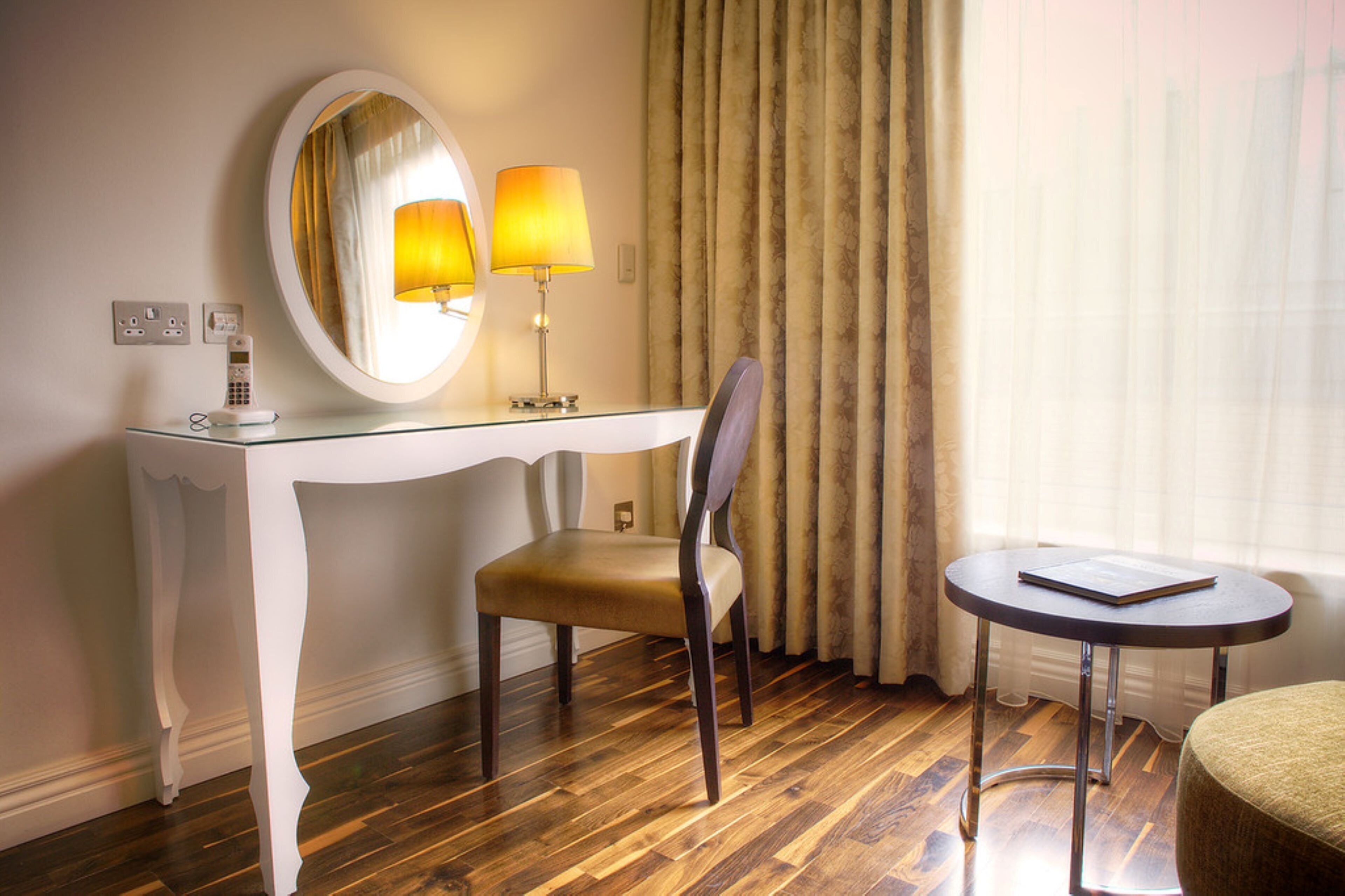 A hotel room with a white console table, a chair, a round table, a mirror, and a lamp.