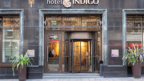 Exterior del Hotel Indigo, con un diseño moderno, que destaca su logo y una entrada de vidrio.