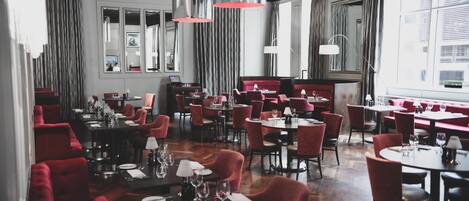 A restaurant with multiple tables and red chairs, large windows, and hanging pendant lights.