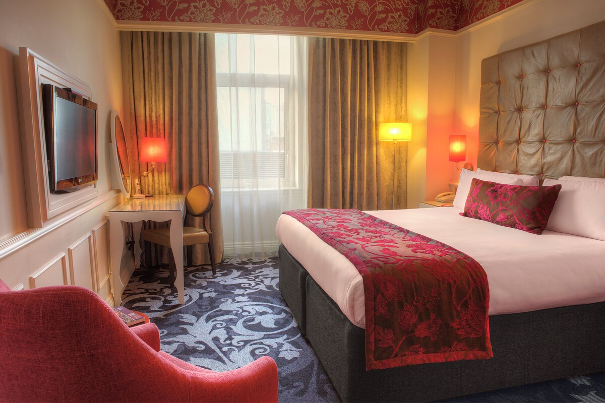 A hotel room with a bed, a desk, a chair, a television, and two lamps.
