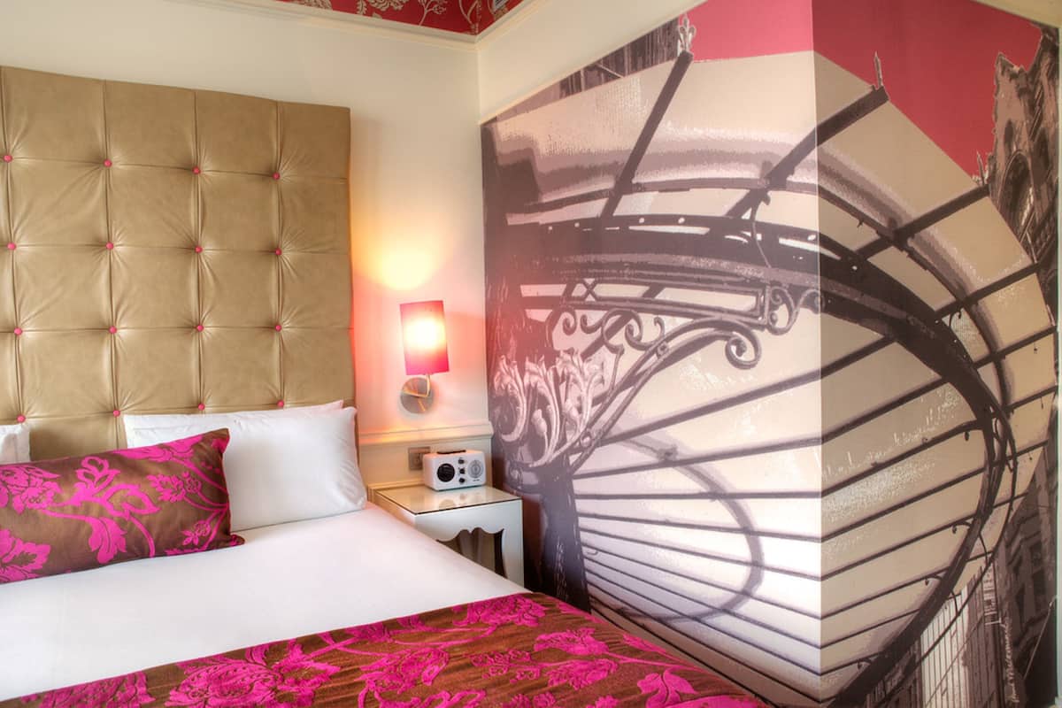 A hotel room with a tufted headboard, a bed with pink floral bedding, a bedside table with a radio, and a large wall mural.