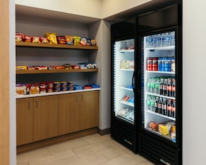 Angolo cottura con scaffali pieni di snack e un frigorifero con acqua in bottiglia e bibite.