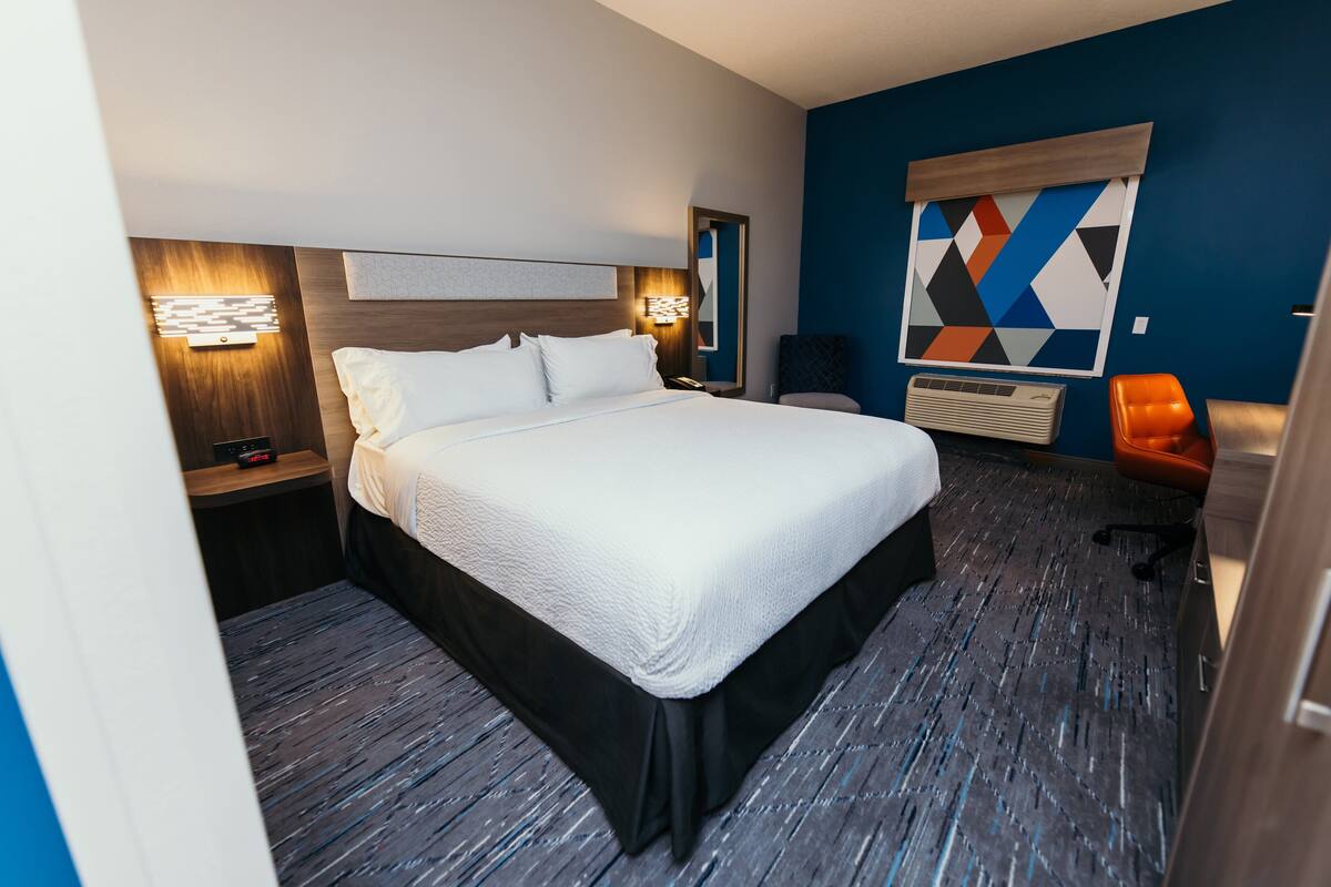 A hotel room with a large bed, a nightstand, a desk, a chair, and a wall-mounted air conditioning unit.