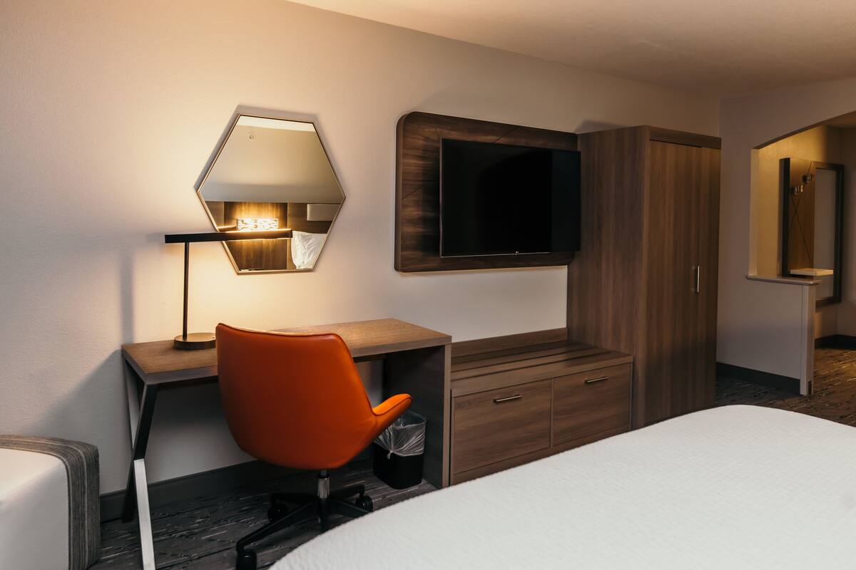 A hotel room with a bed, a desk with a chair, a TV mounted on the wall, and a wooden wardrobe.