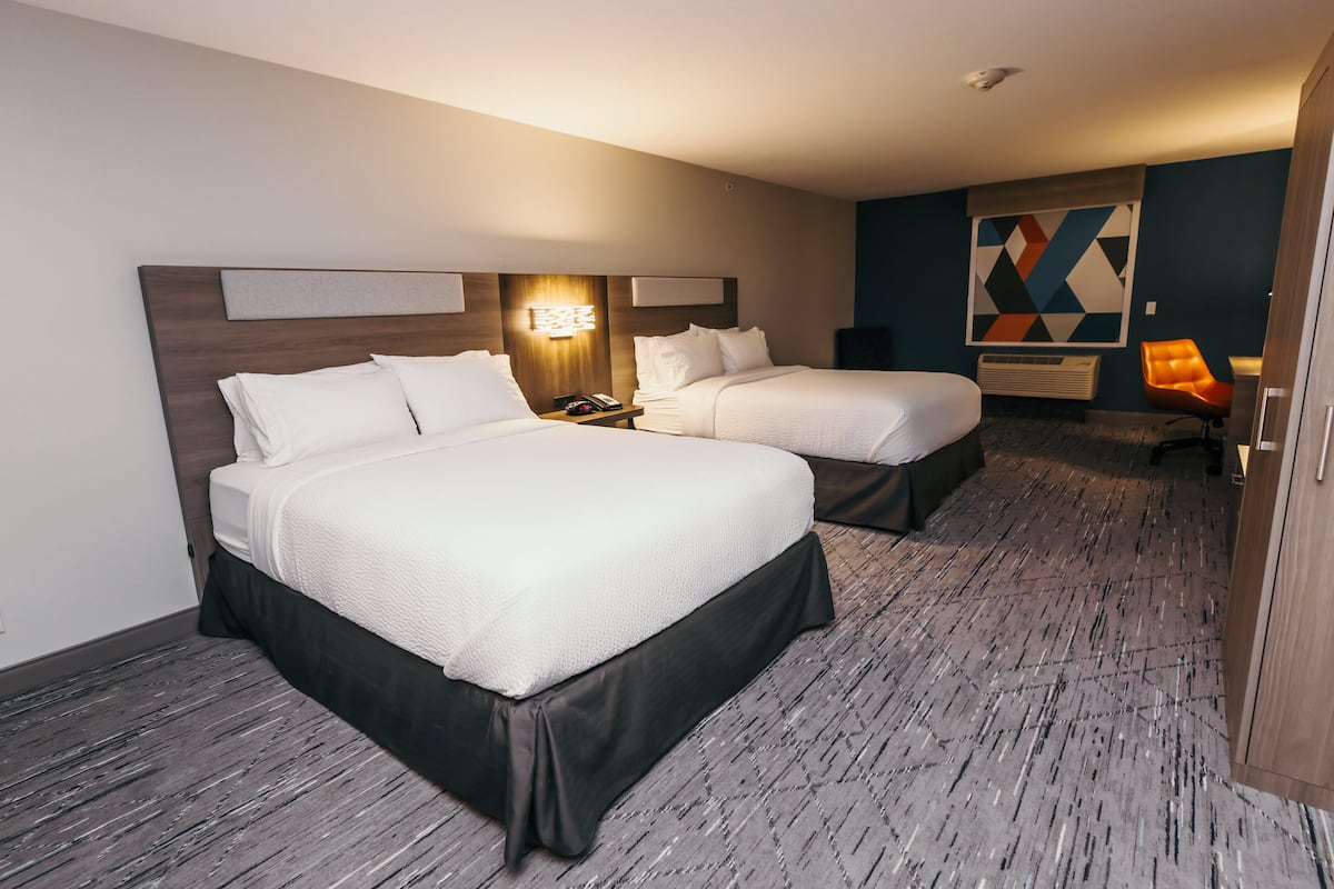 A hotel room with two beds, a nightstand, a lamp, a chair, and a geometric wall decoration.