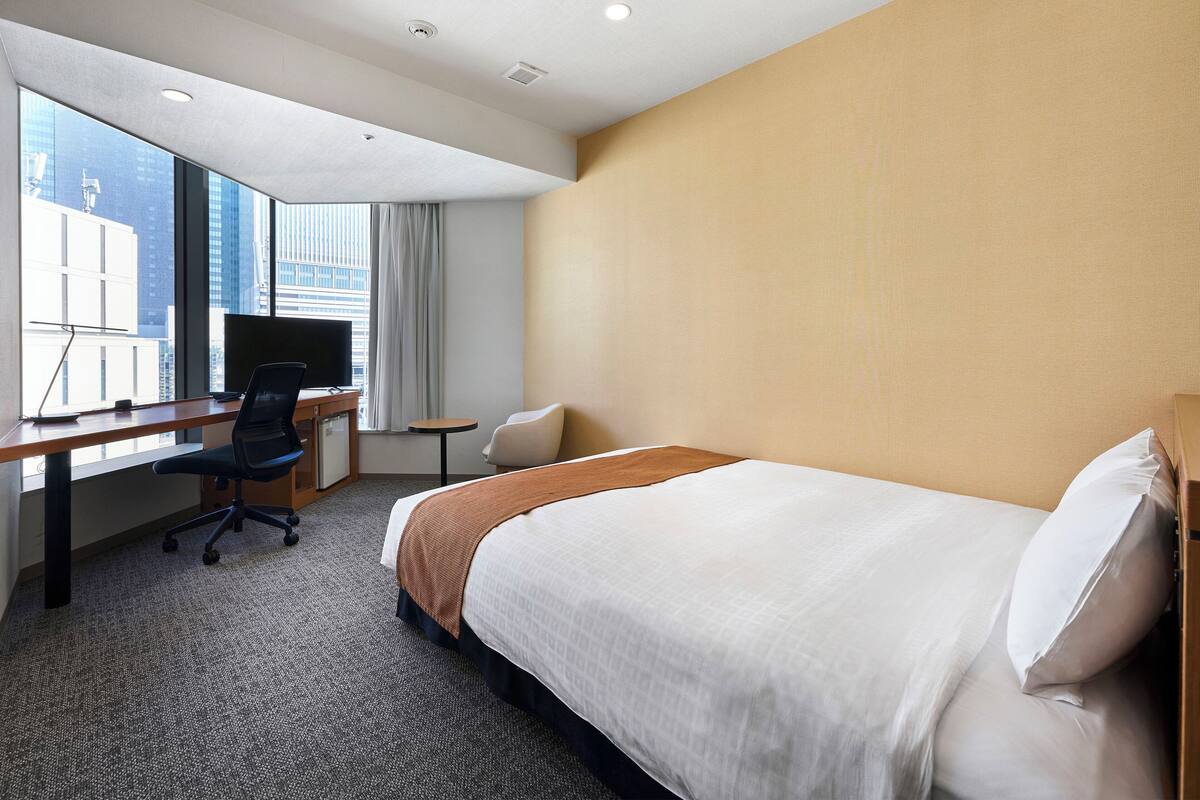 A hotel room with a bed, a desk with a chair, a TV, and a window with curtains.