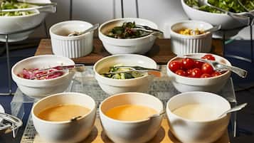 Mesa de buffet com diversos pratos em recipientes, incluindo saladas, legumes e possivelmente carne.