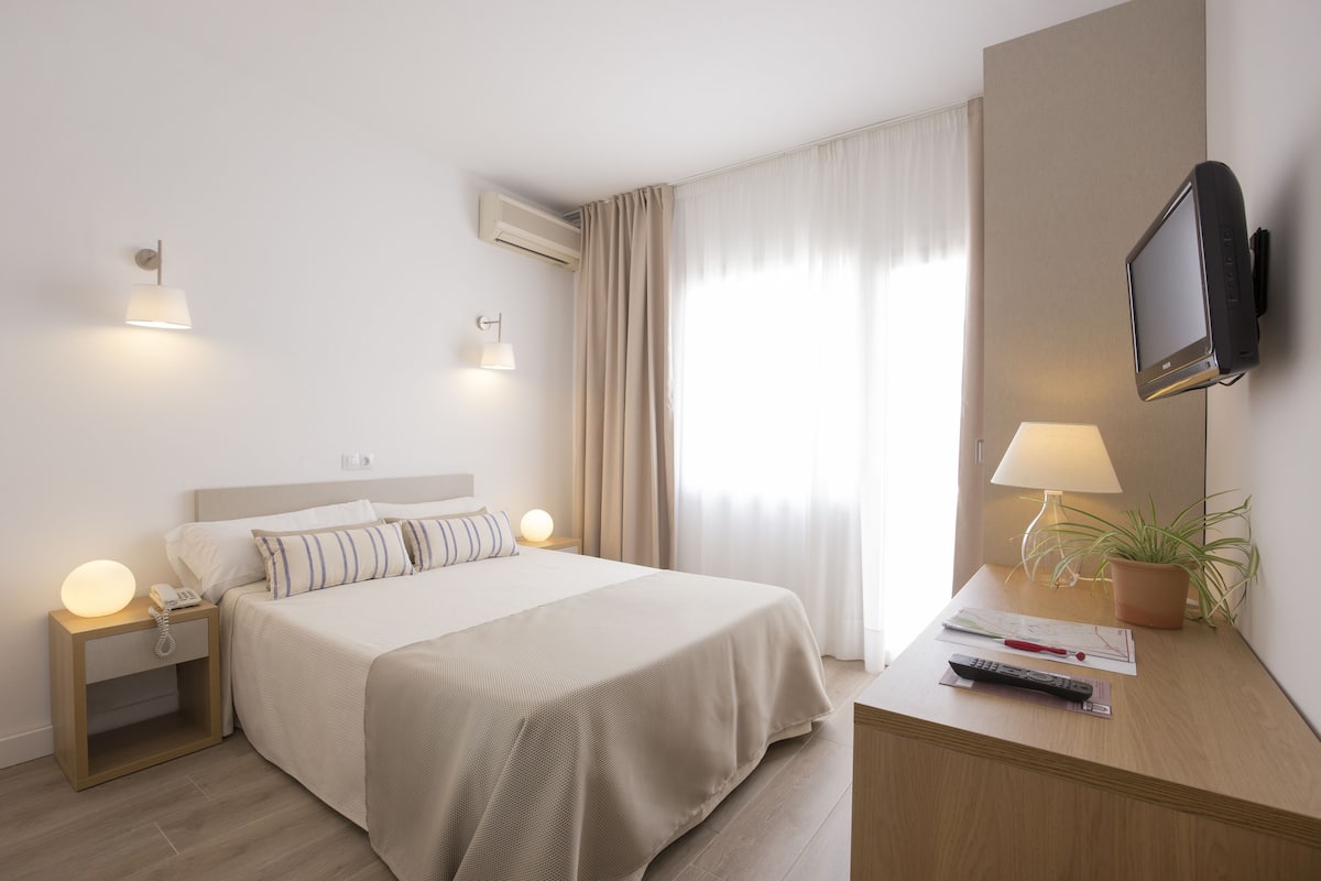 Quarto de hotel com cama, abajures, TV fixada na parede, escrivaninha com luminária e planta em vaso.