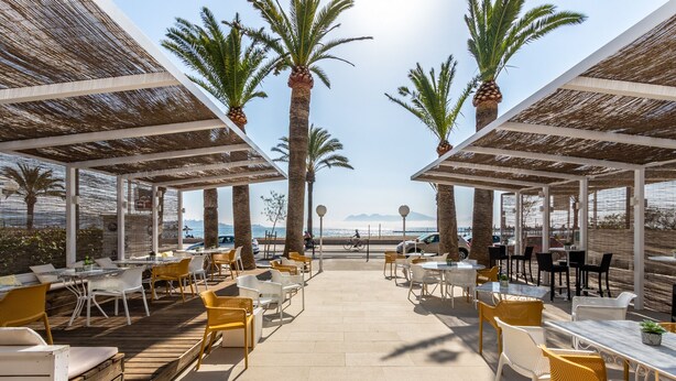 An outdoor café with tables and chairs, palm trees, and a view of the sea. -  Hotel Cabot Romantic - Adults Only (Pollenca)