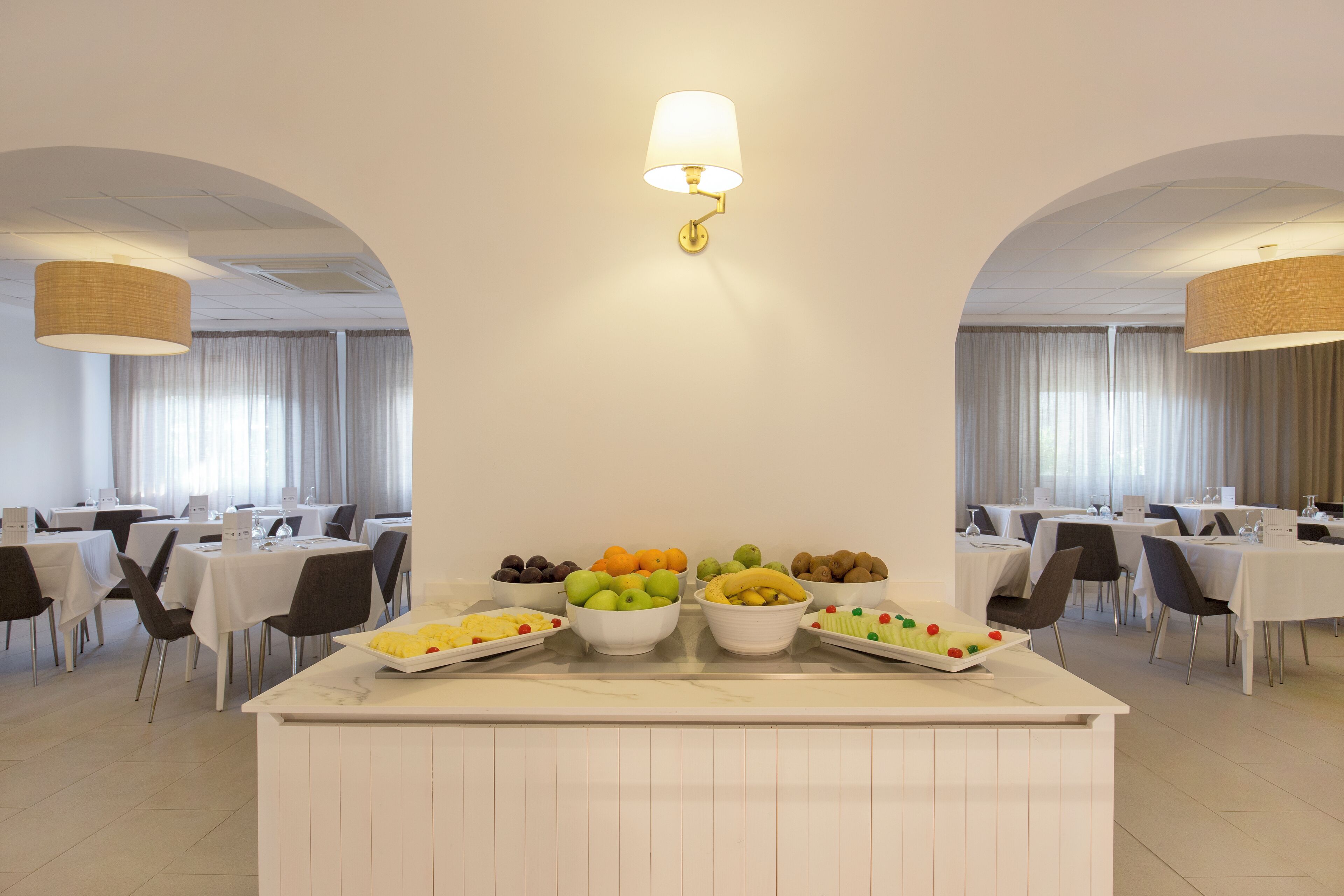 Een eetruimte met witte tafels en stoelen, een centraal fruitplateau en een plafondlamp.