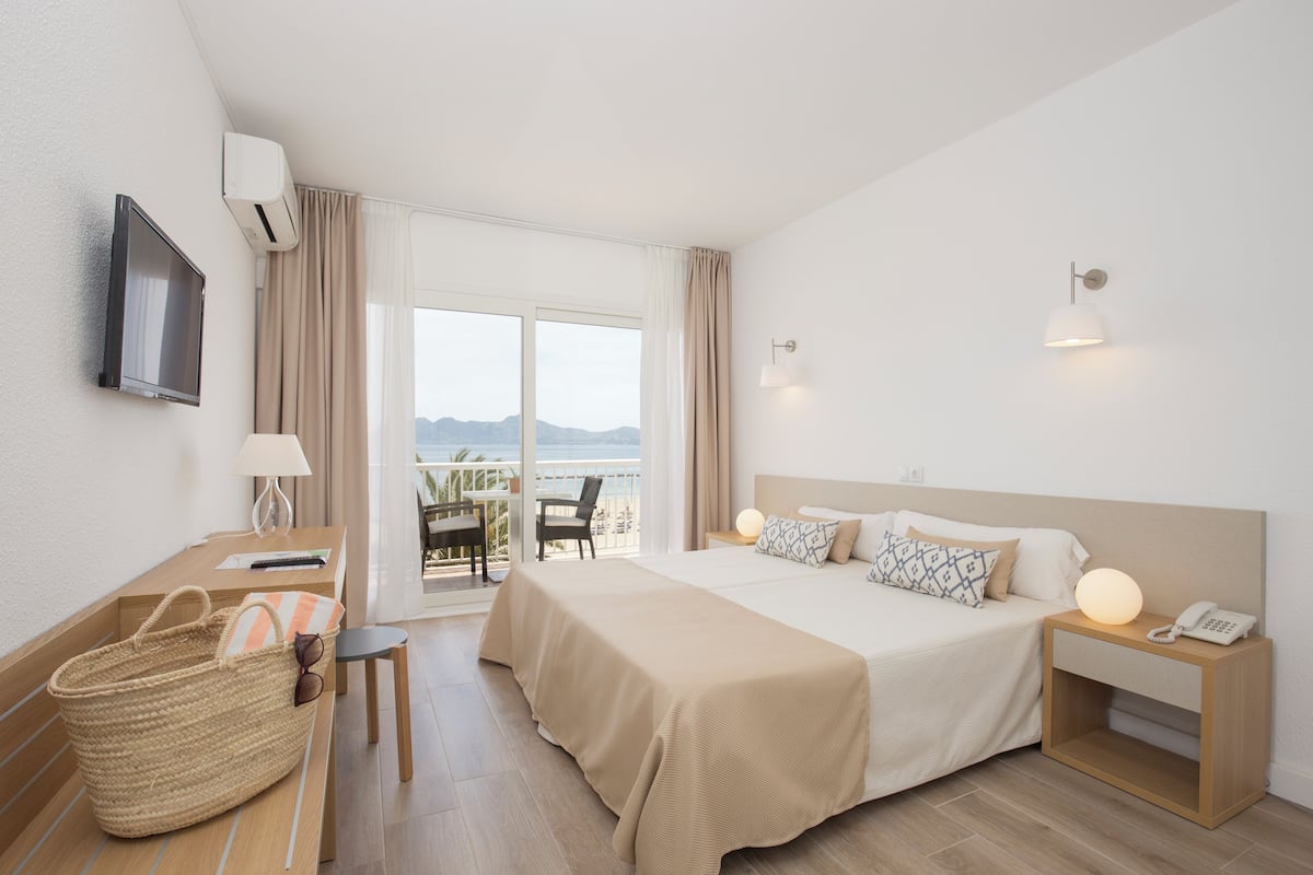Quarto de hotel com cama, mesa de cabeceira, abajur e vista para o mar.