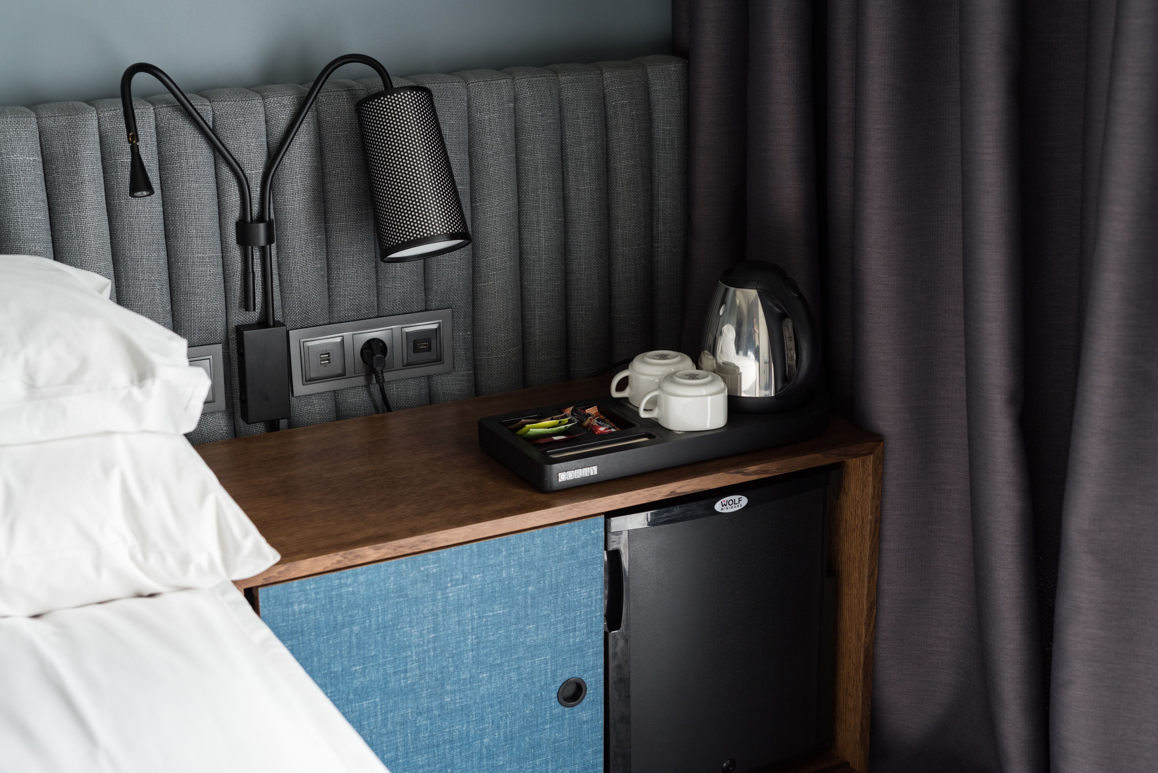 A hotel room with a bed, a bedside table with a lamp, a coffee maker, and a small refrigerator.