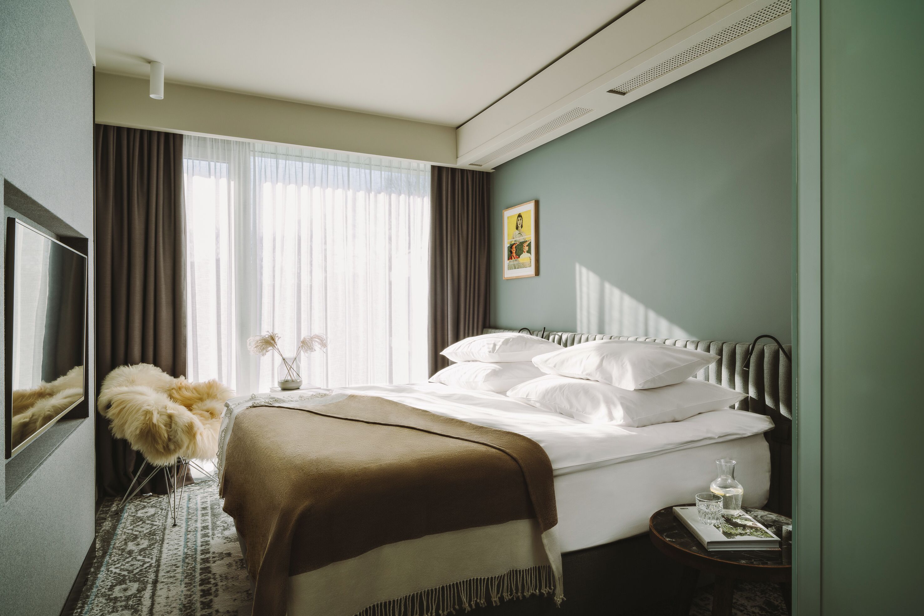 Una camera d'albergo con un letto, una televisione, un tavolino con un libro e una poltrona decorativa.