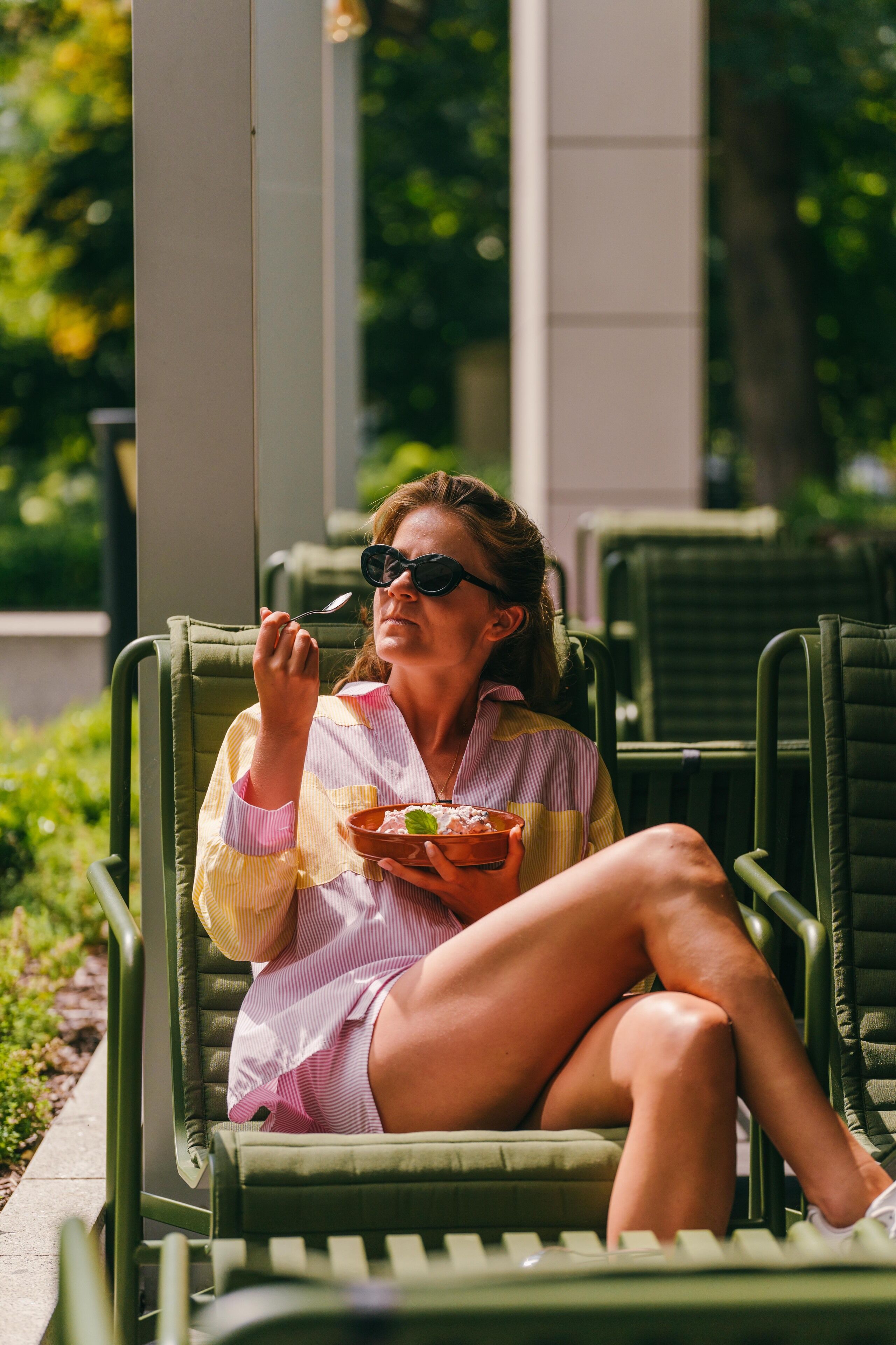A person sitting on a green lounge chair outdoors, holding a bowl and spoon, with greenery in the background.