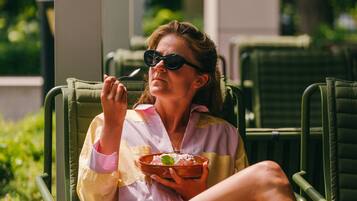 A person sitting on a green lounge chair outdoors, holding a bowl and spoon, with greenery in the background.