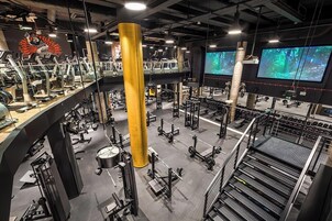 Una palestra su più livelli con diverse attrezzature per esercizi, un’imponente colonna centrale e numerosi schermi che mostrano scorci naturali.