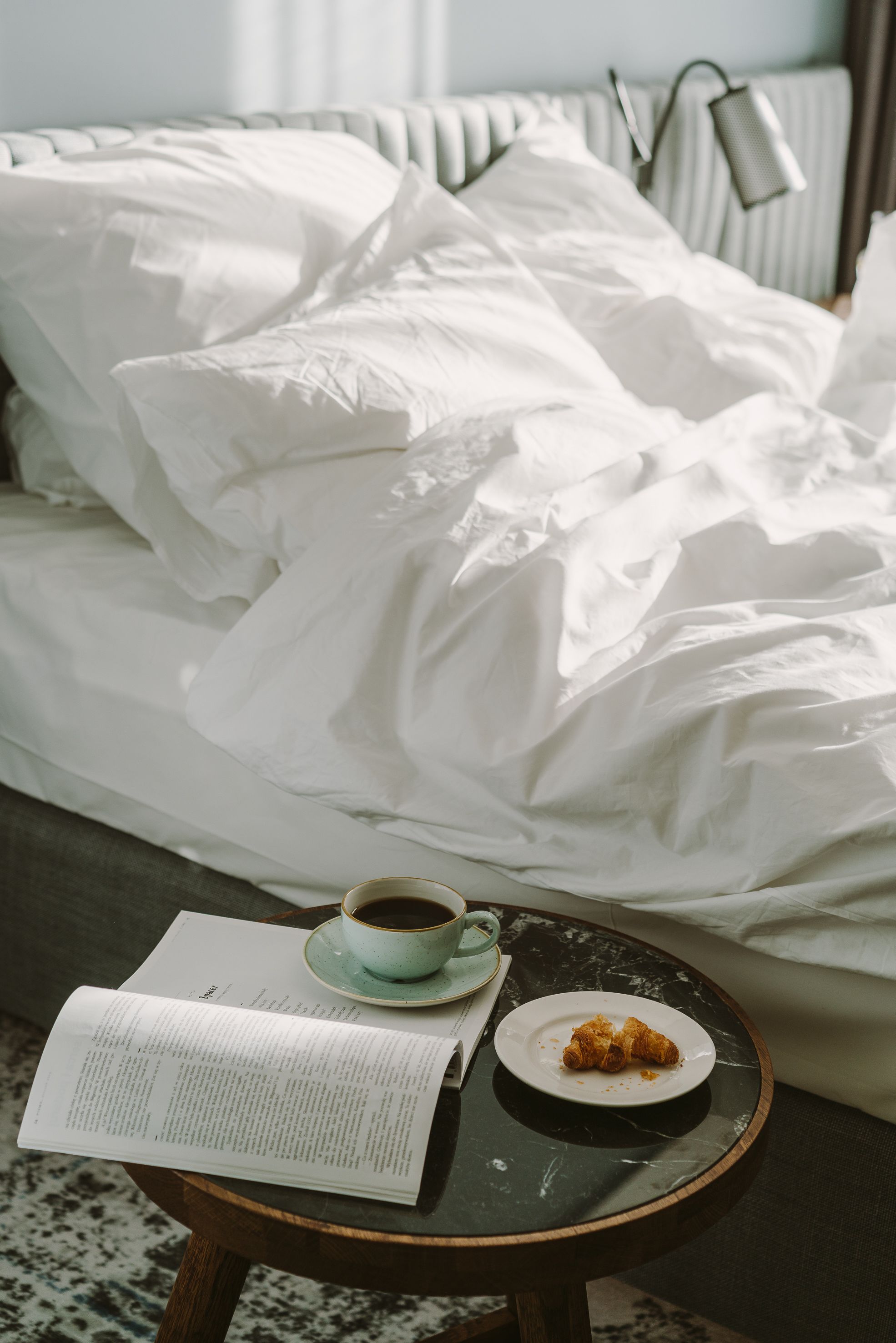 A neatly made bed with white linens, a cup of coffee on a saucer, a plate with a pastry, and an open menu on a small table.