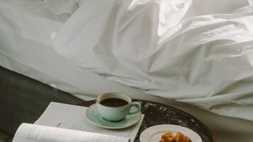 A neatly made bed with white linens, a cup of coffee on a saucer, a plate with a pastry, and an open menu on a small table.