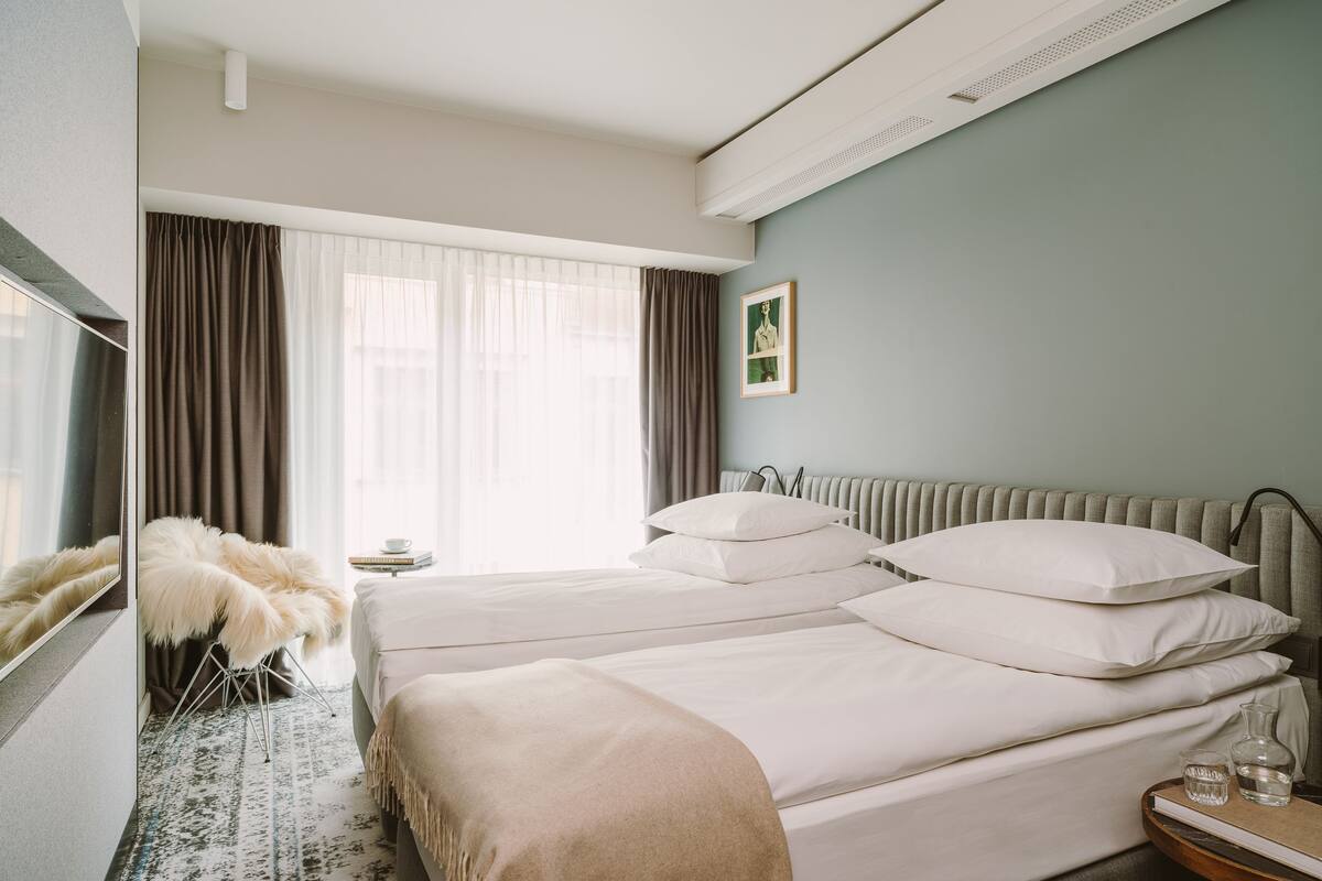 Une chambre d’hôtel avec deux lits, une grande fenêtre avec des rideaux, un radiateur, un tableau encadré au mur et une petite table sur laquelle se trouvent un vase et un livre.