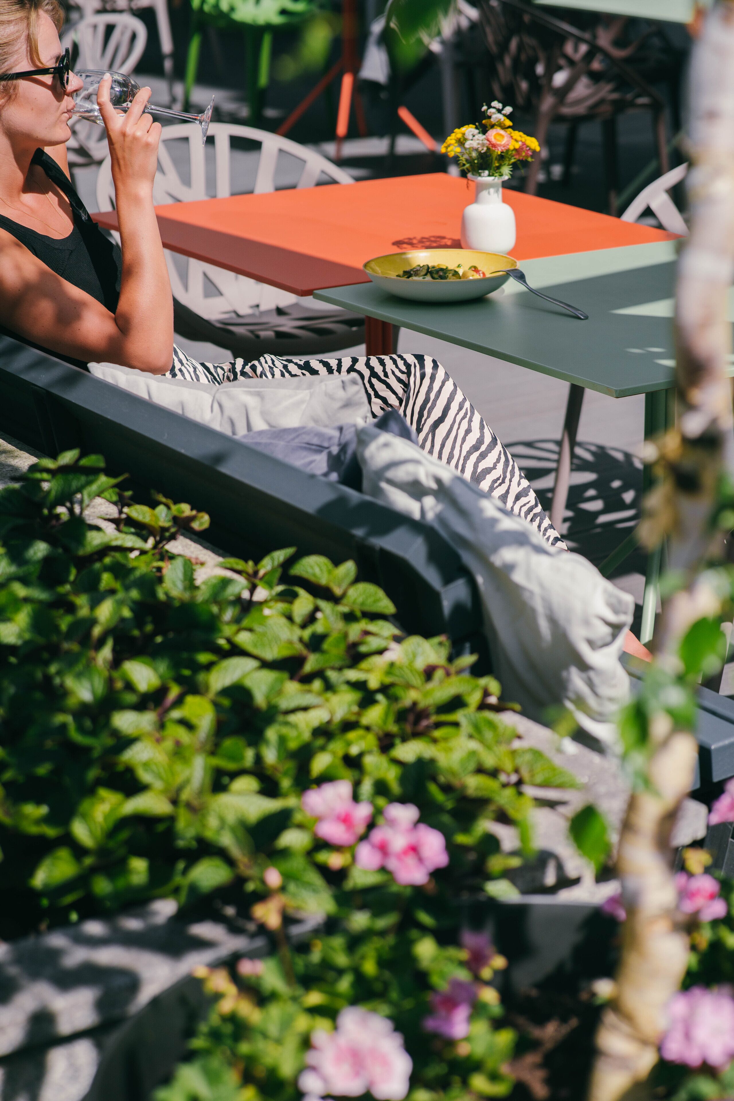 Una persona recostada en un banco acolchado al aire libre, con una mesa y un florero cerca.