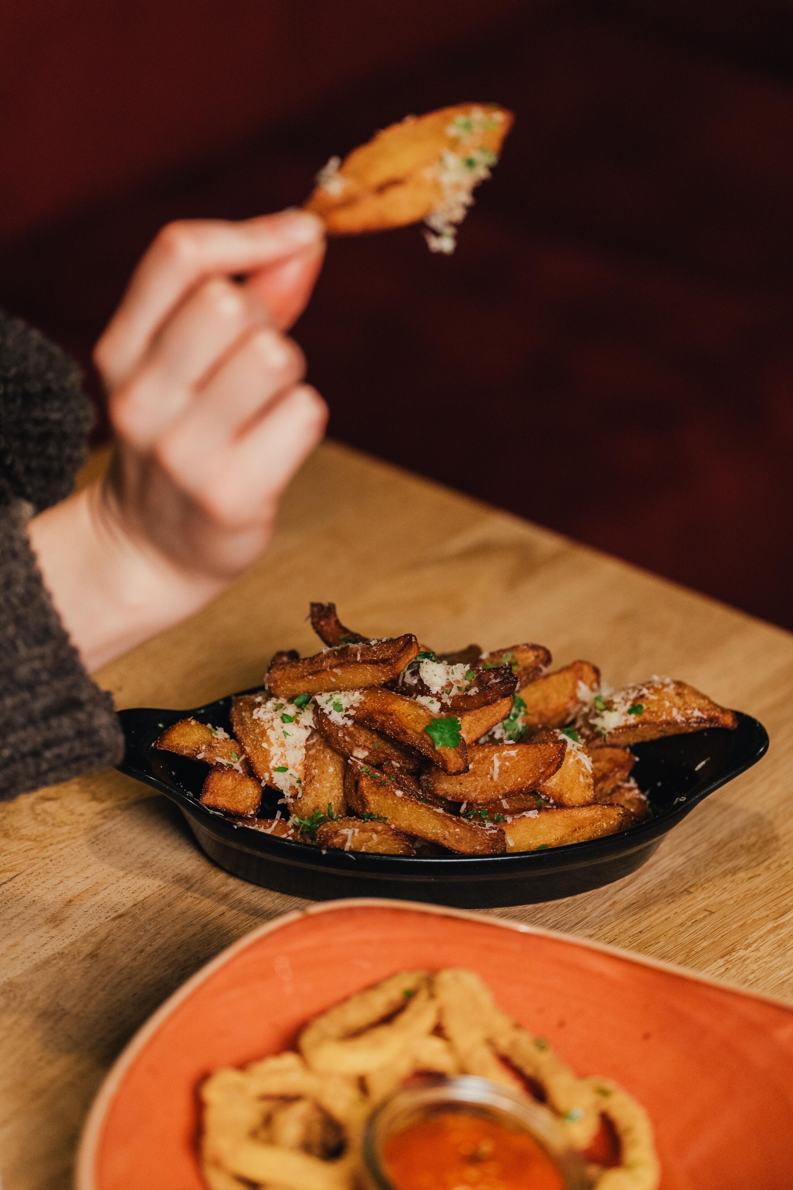 A person holding a piece of food over a black skillet filled with fries, with a bowl of onion rings and dipping sauce in the foreground.