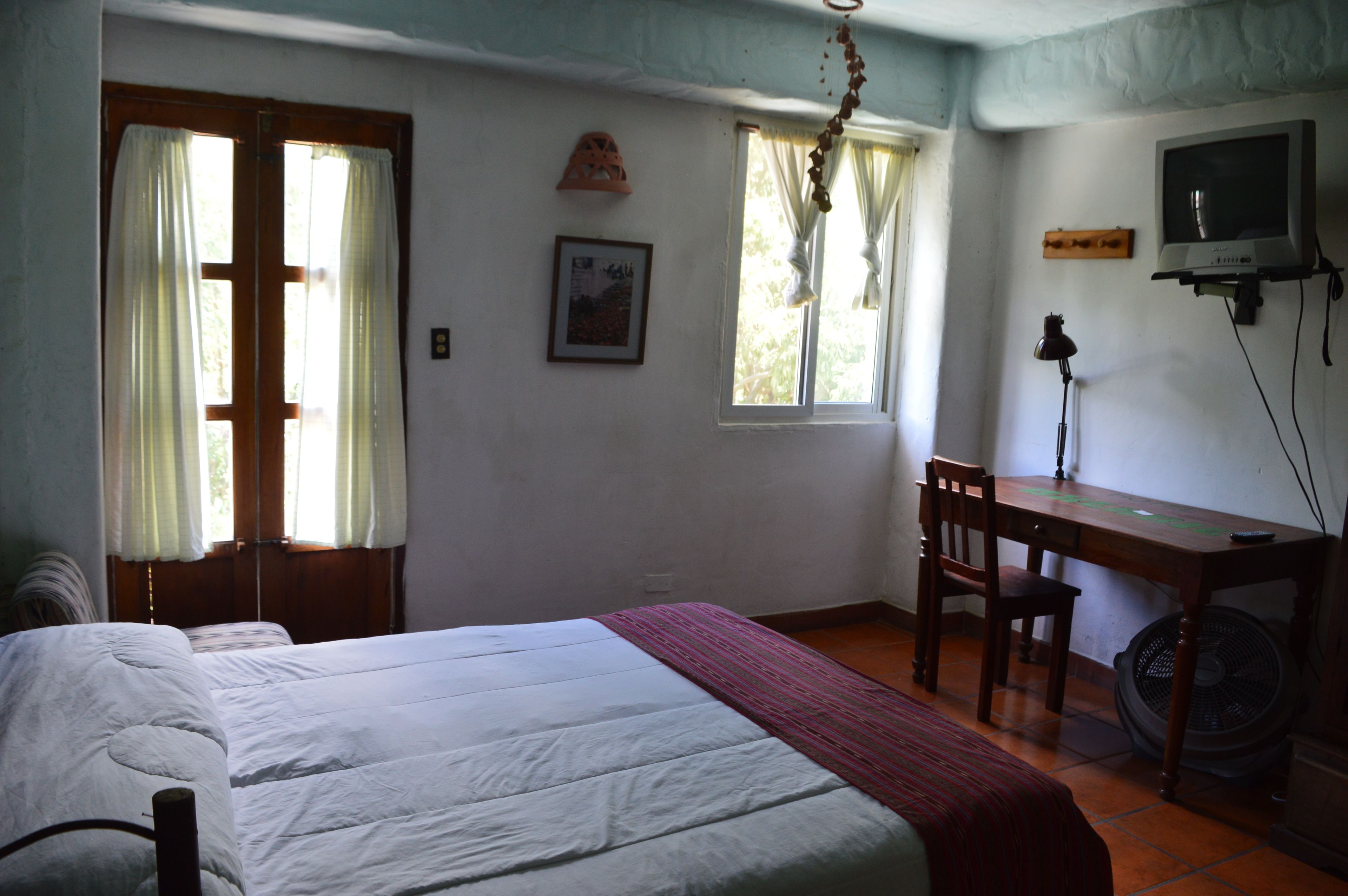 Une chambre à coucher avec un lit, un bureau et une chaise. Il y a une fenêtre avec des rideaux, un tableau accroché au mur et un téléviseur fixé au mur.