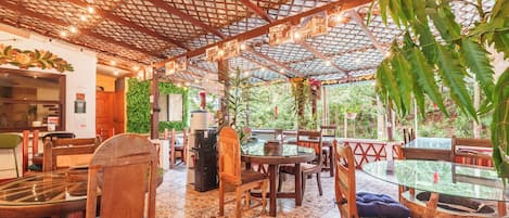 Un área de comedor al aire libre con mesas y sillas de madera, suelo de baldosas y techo con diseño de cuadrícula.