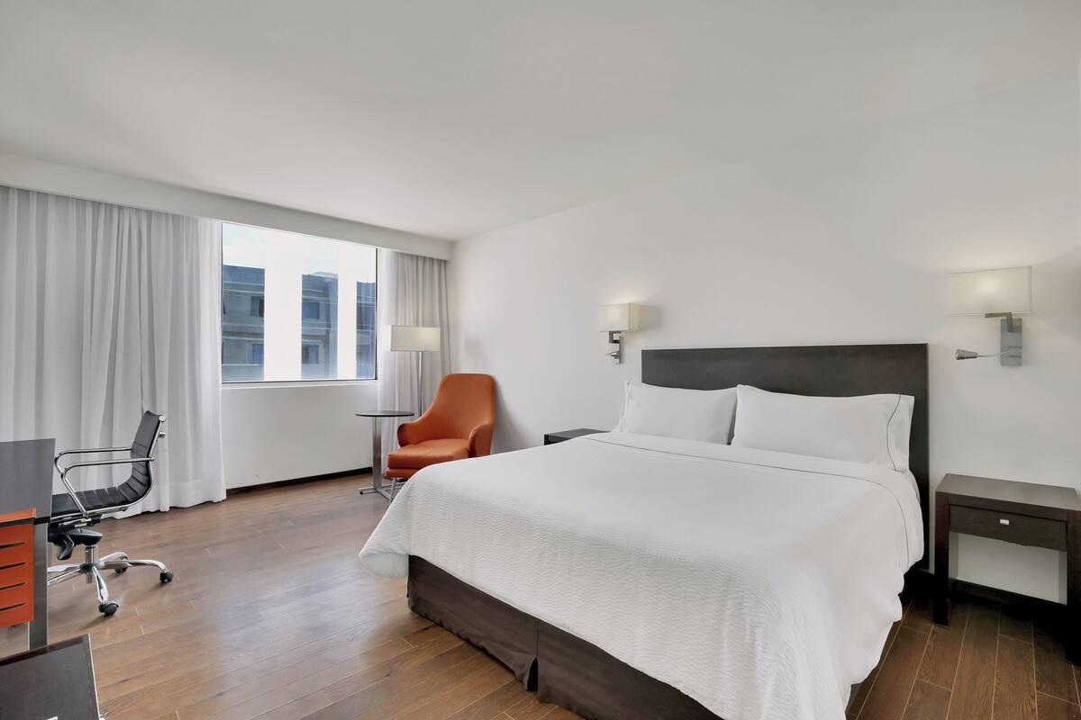 A hotel room with a large bed, a desk with a chair, a small table, and a window with curtains.