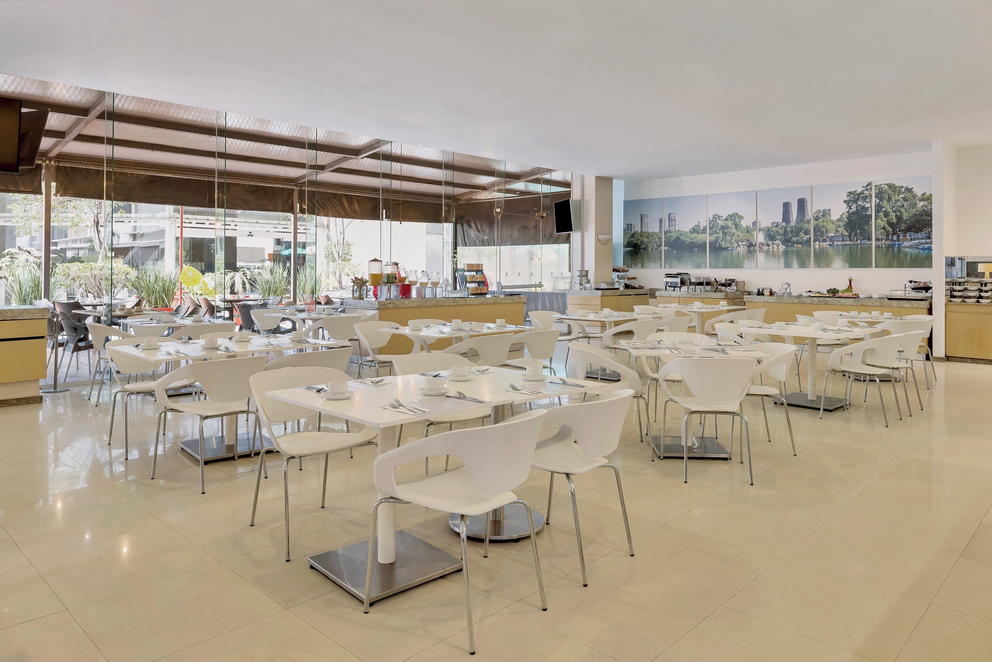 A spacious indoor dining area with white tables and chairs, large windows with a view of buildings and trees, and a mural of a lake and city skyline.